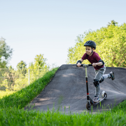 Small child in red t-shirt, grey pants and a helmet riding a scooter on pump track surrounded by green grass