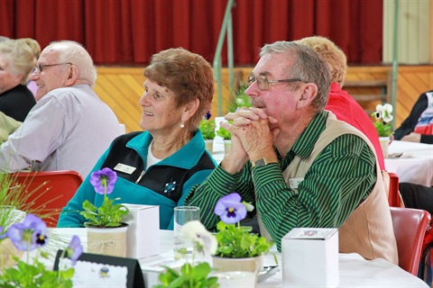 Man and woman wearing volunteer badges, seated at a table looking to the front of a the room.
